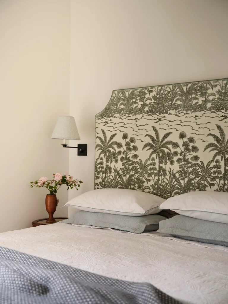 Cozy bedroom with tropical-patterned headboard, white pillows, white bedding and bed sheets, blue blanket and a wooden side table with a vase of pink flowers.