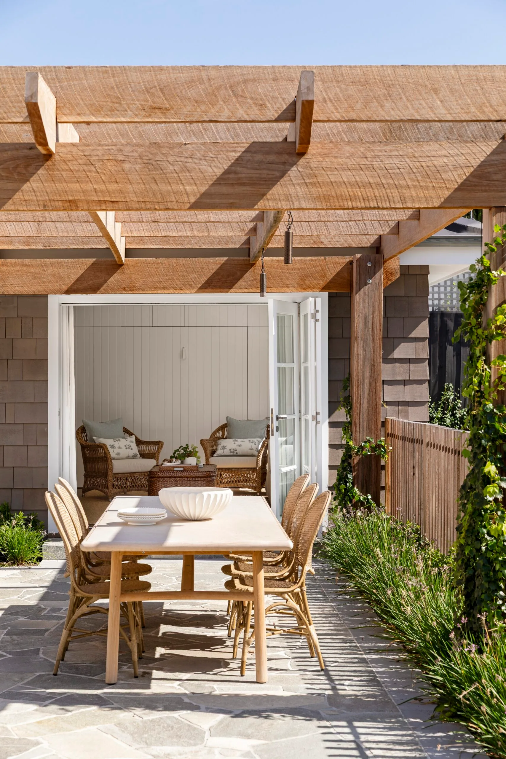 Outdoor patio with timber pergola, dining table, and rattan chairs; open doors reveal wicker armchairs and cushions inside.