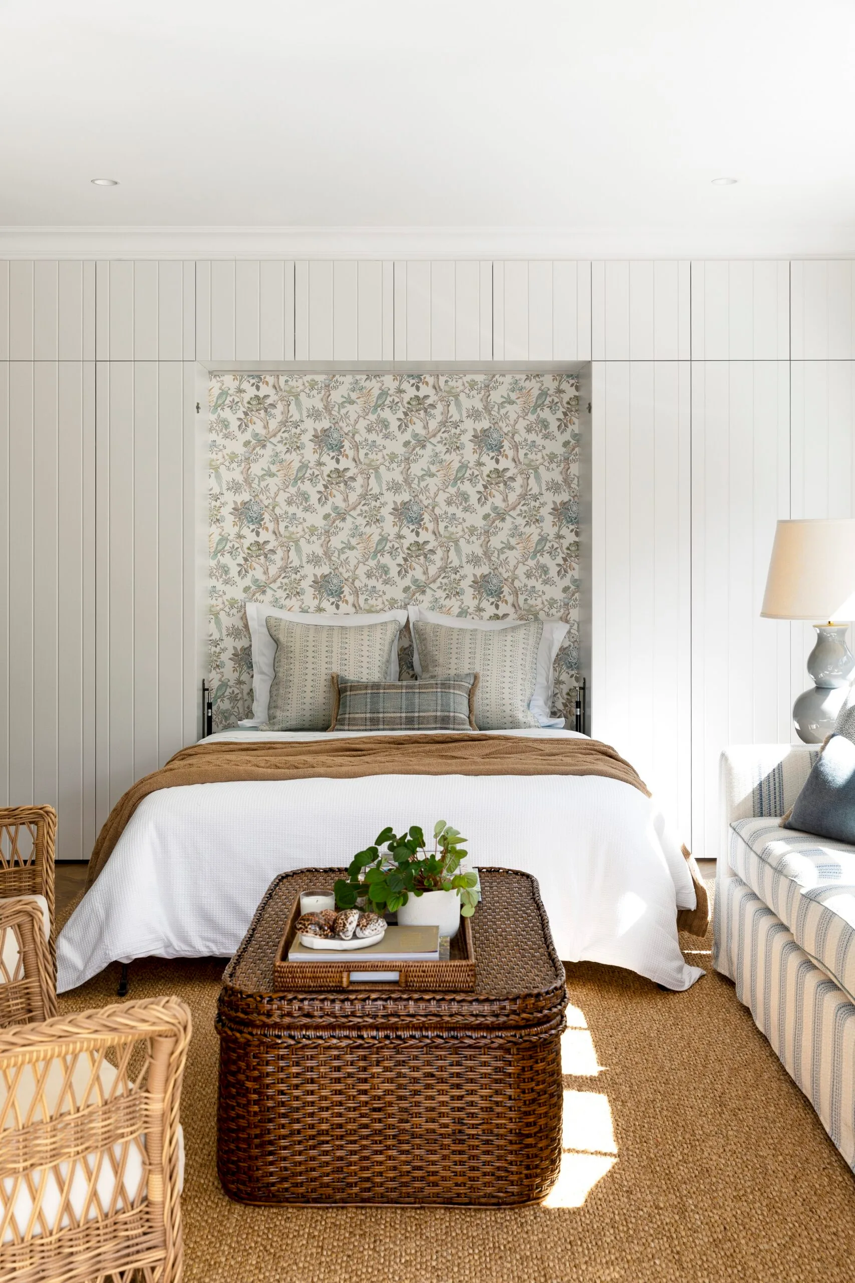 Cosy bedroom in a Cape Cod style home. Built-in cabinetry with no handles surrounds the bed. The bed is positioned in an alcove with floral wallpaper. In the foreground, wicker furniture, and earthy tones. This is all atop a sisal rug.