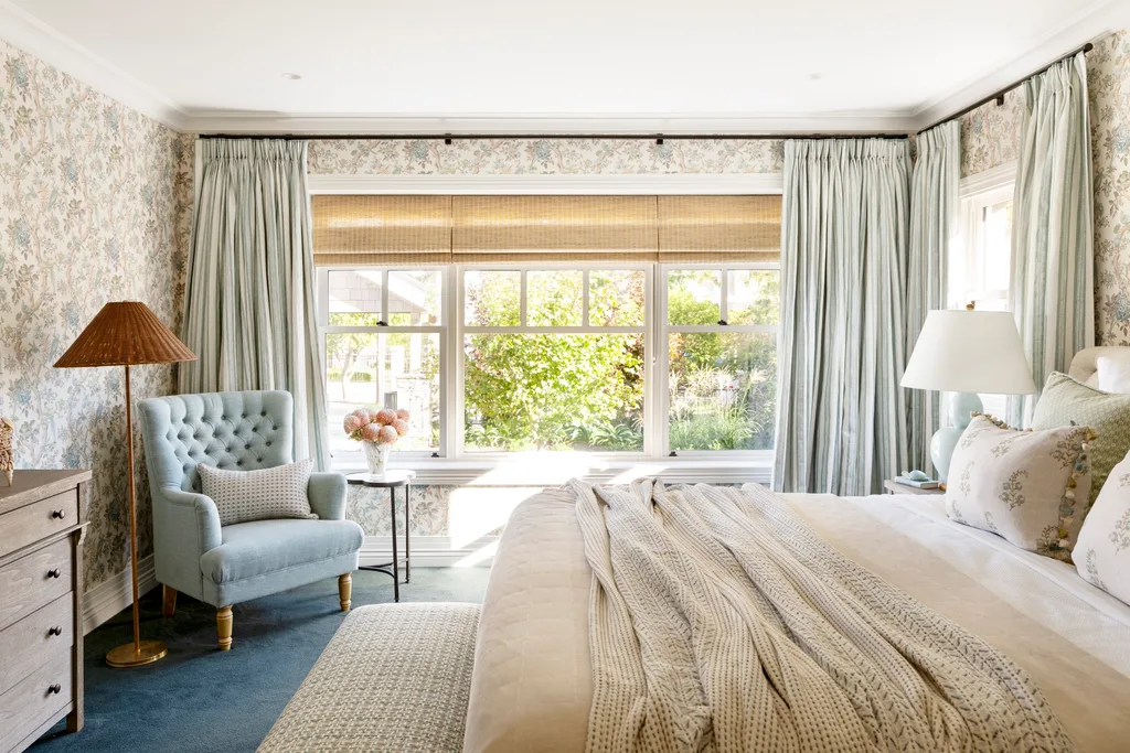 Cozy bedroom with large window, floral wallpaper, blue armchair, side table with flowers, and a bed with a knit blanket.