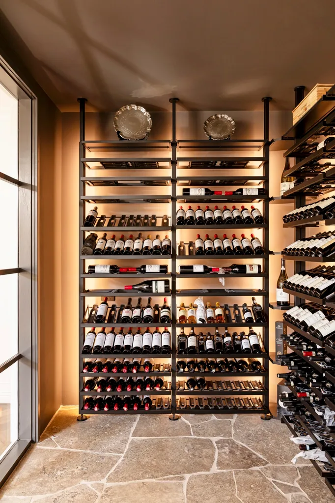 A cellar with paved stone flooring, black shelves with bottles of wine leading from floor to ceiling.