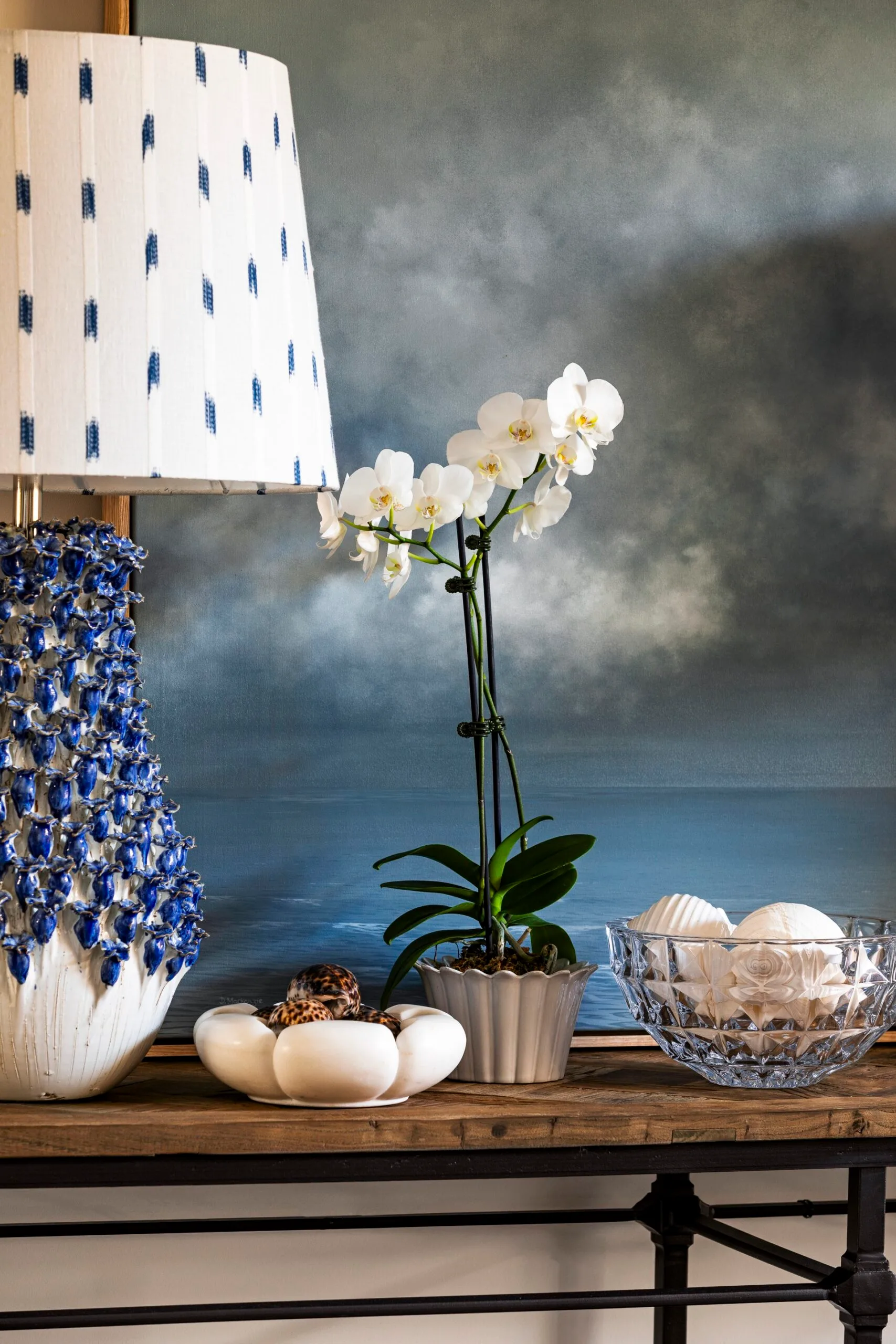 White orchids and seashells displayed in a crystal bowl on a small timber console table with black steel legs. A textured blue and white lamp base and a patterned lampshade are on the table, against a cloudy sky painting.