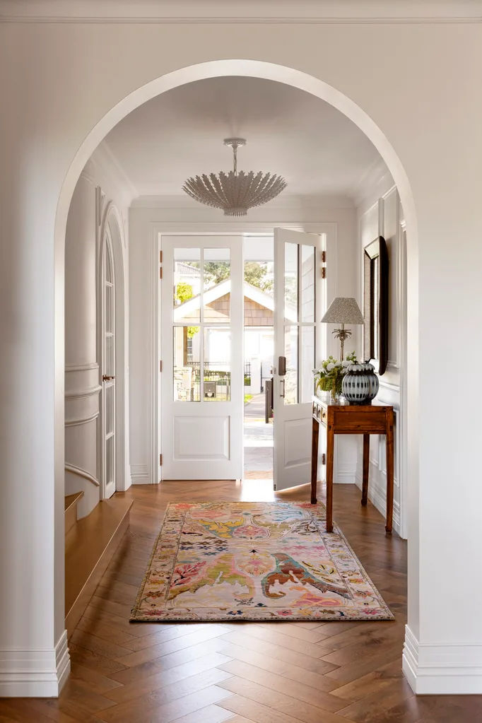 Elegant hallway in a Cape Cod style home with arch, patterned rug, table with lamp, and open French doors leading outside. Timber floors and ornate skirting boards add heritage charm.