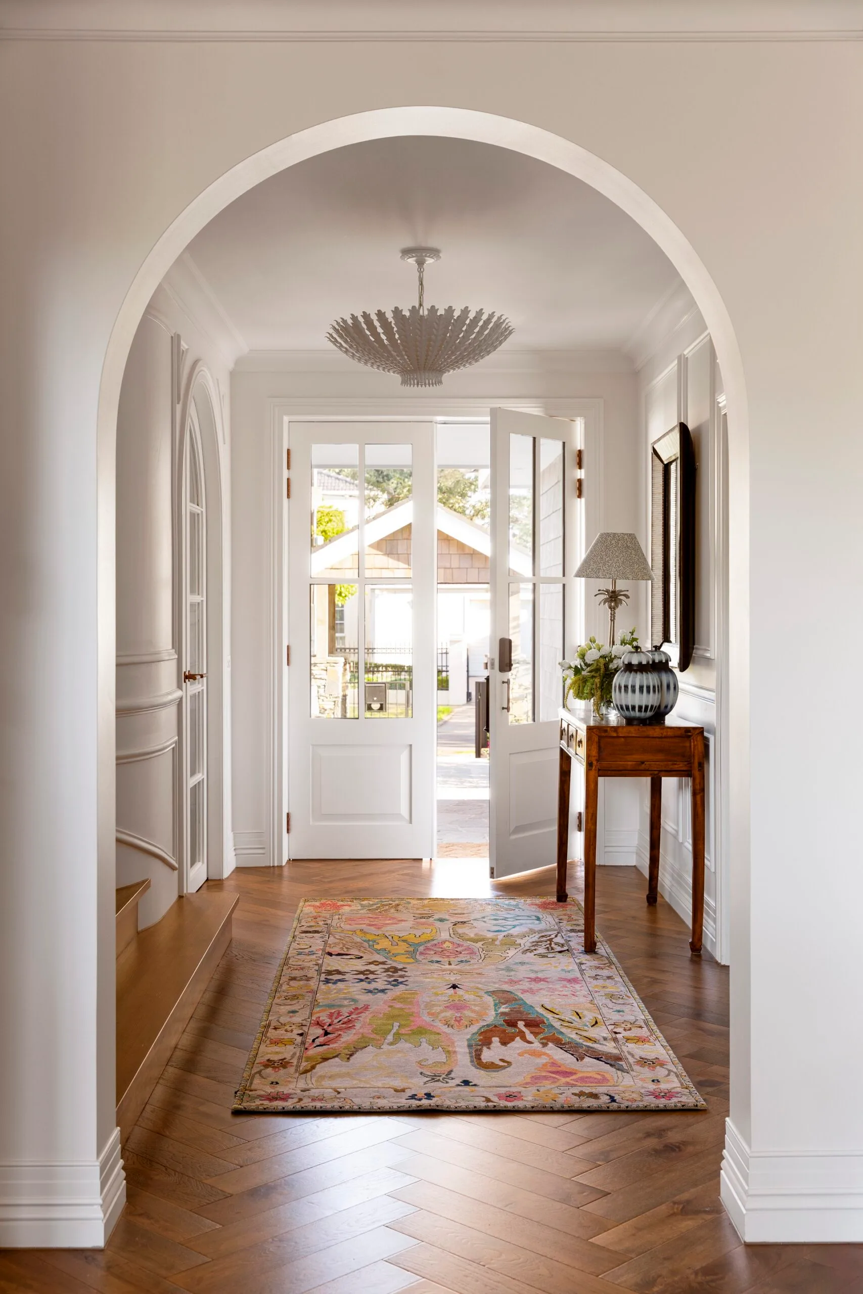 Elegant hallway in a Cape Cod style home with arch, patterned rug, table with lamp, and open French doors leading outside. Timber floors and ornate skirting boards add heritage charm.