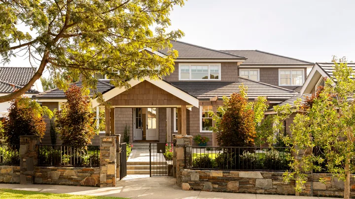 A Cape Cod style home exterior, with lots of trees and bushes in the front garden. THe home is two stories and features white window frames and brown cladding on the walls. A path leads to the front door and stones comprise a small fence leading to a black steel gate.
