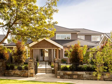 A Cape Cod style home exterior, with lots of trees and bushes in the front garden. THe home is two stories and features white window frames and brown cladding on the walls. A path leads to the front door and stones comprise a small fence leading to a black steel gate.