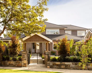 A Cape Cod style home exterior, with lots of trees and bushes in the front garden. THe home is two stories and features white window frames and brown cladding on the walls. A path leads to the front door and stones comprise a small fence leading to a black steel gate.