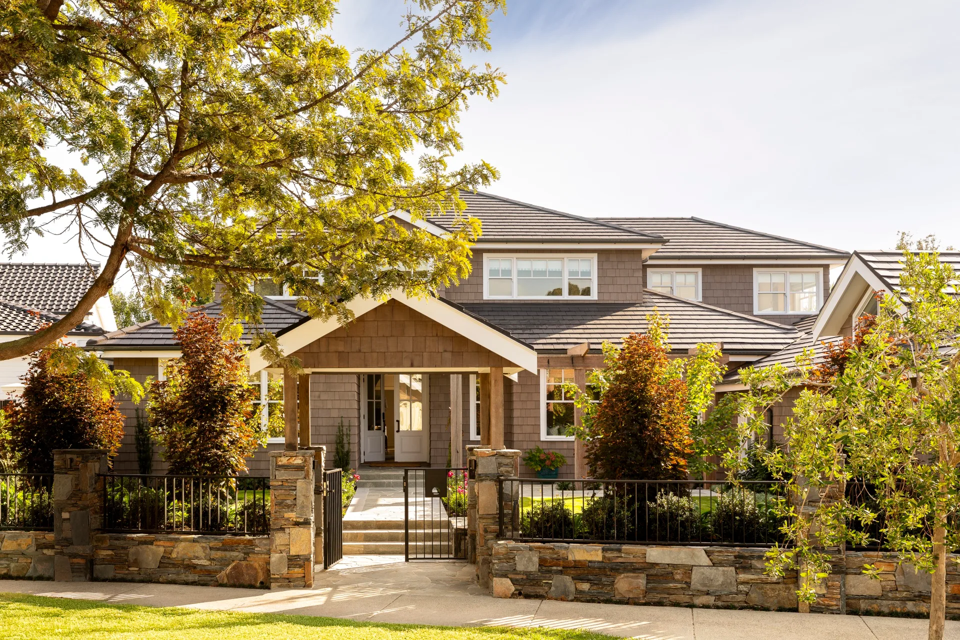 A Cape Cod style home exterior, with lots of trees and bushes in the front garden. THe home is two stories and features white window frames and brown cladding on the walls. A path leads to the front door and stones comprise a small fence leading to a black steel gate.