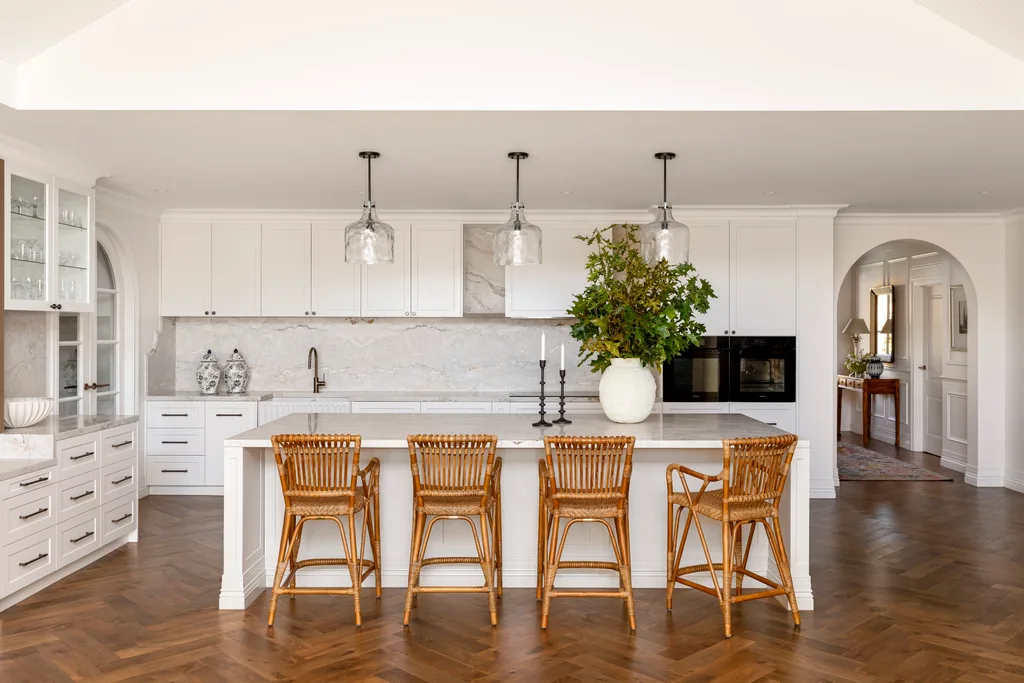 Elegant kitchen with white cabinets, marble splashback and benchtop, timber and woven bar stools tucked under the central island and three pendant lights positioned above. This open plan kitchen is in a Cape Cod style home.