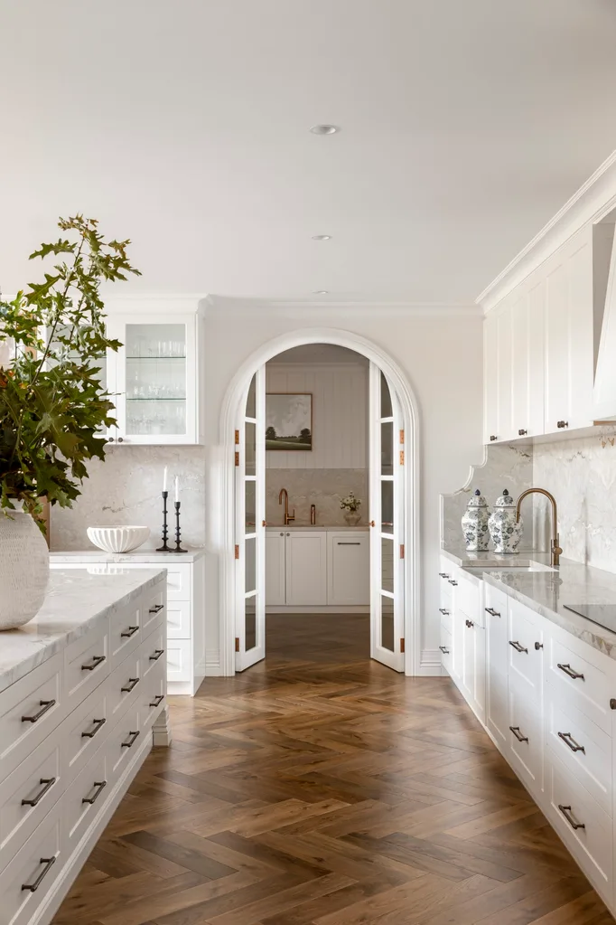 Elegant white kitchen with arched French doors leading to a butler's pantry, marble countertops and splashback, and herringbone timber floors. A large vessel with a leafy green plant is on the kitchen island.