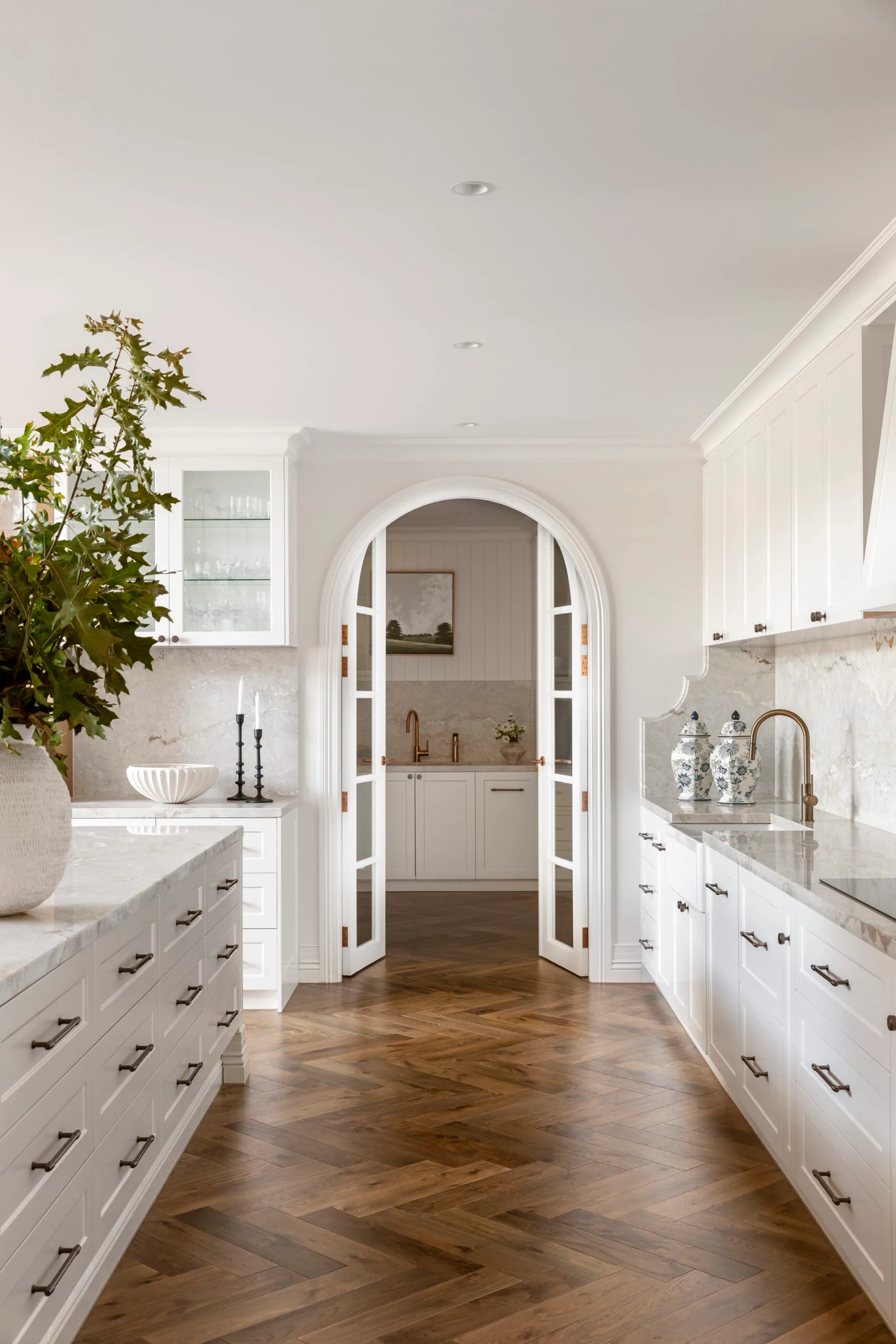 Elegant white kitchen with arched French doors leading to a butler's pantry, marble countertops and splashback, and herringbone timber floors. A large vessel with a leafy green plant is on the kitchen island.