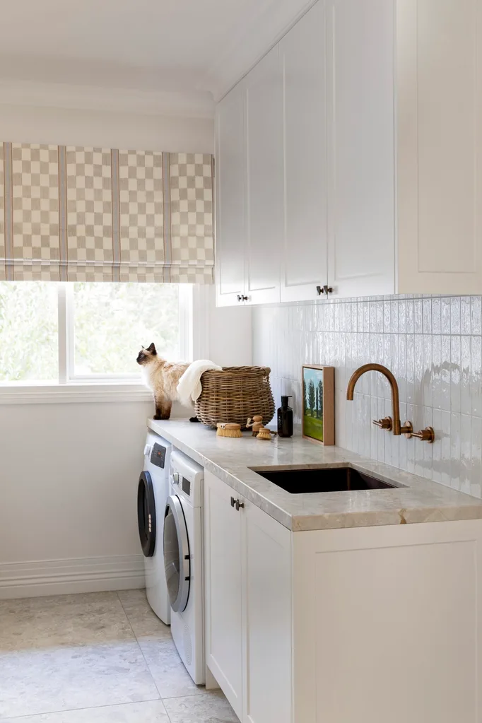 A cat on a laundry room countertop, next to a woven basket, in a bright space with a washer, sink, and checkered blinds. Brass hardware is attached to the white tiled splashback.