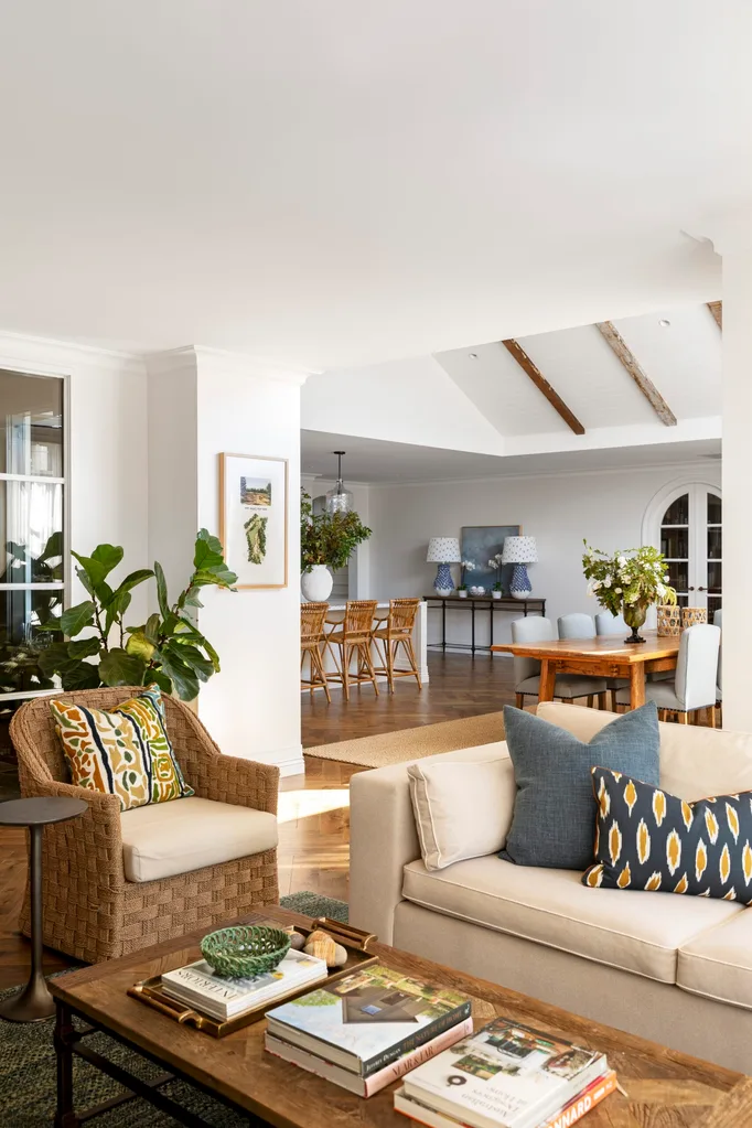 Living room with wicker armchair, patterned cushions on cream toned sofa, coffee table books, and open view to dining area with vaulted ceilings. This open plan space is in a Cape Cod style home.