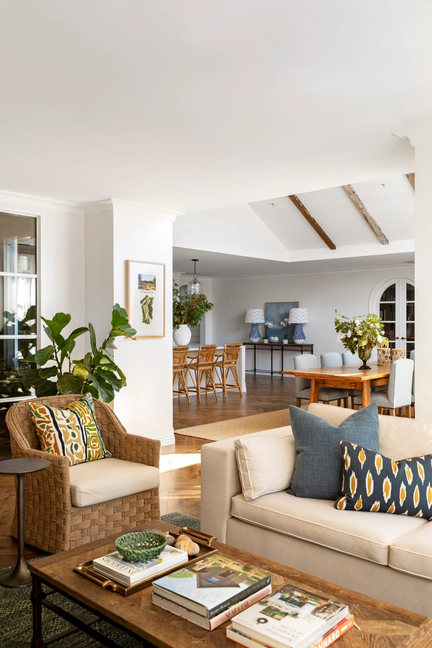 Living room with wicker armchair, patterned cushions on cream toned sofa, coffee table books, and open view to dining area with vaulted ceilings. This open plan space is in a Cape Cod style home.