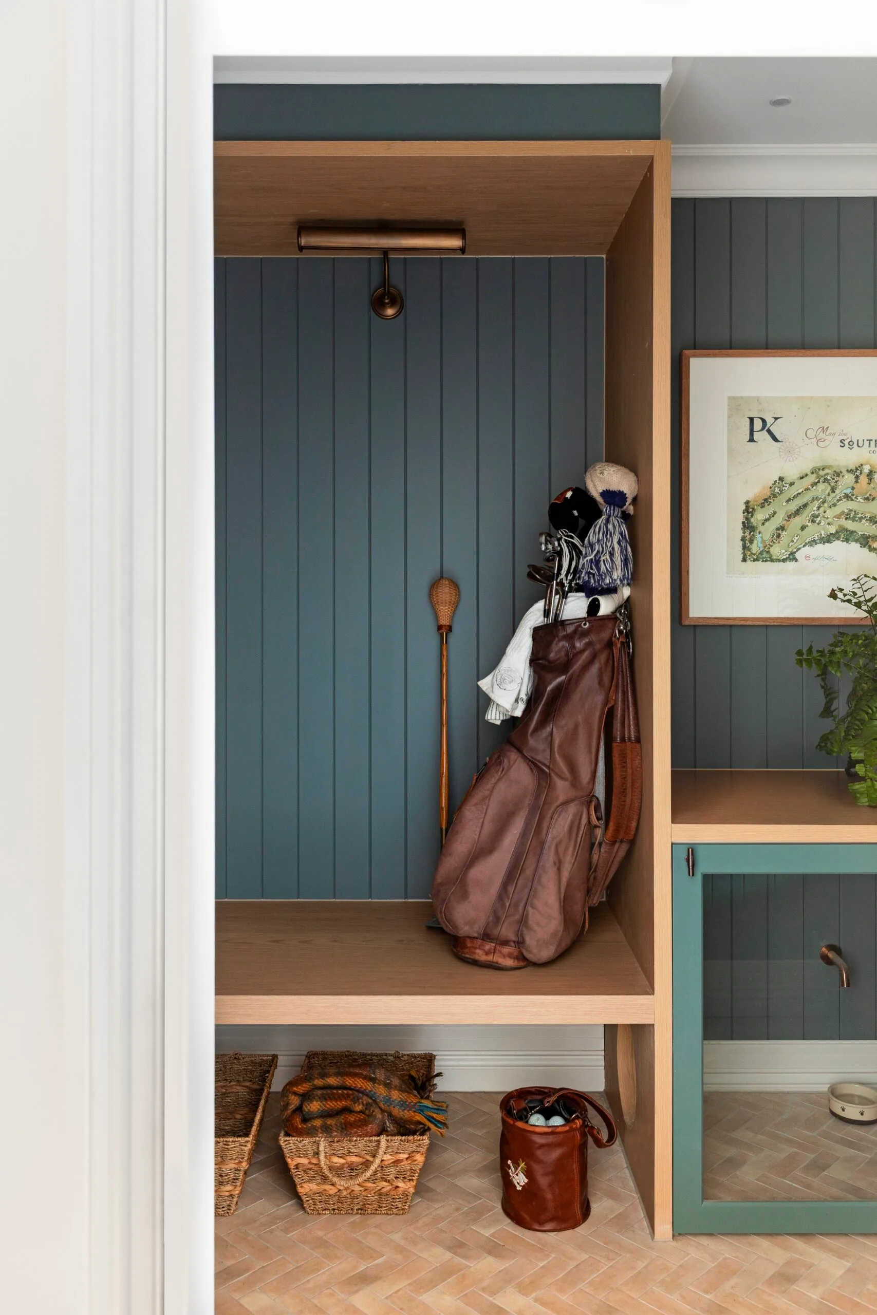 A mudroom within a Cape Cod style home. Golf bag with clubs and baskets in a wooden cubby against blue paneled wall, next to a framed map.