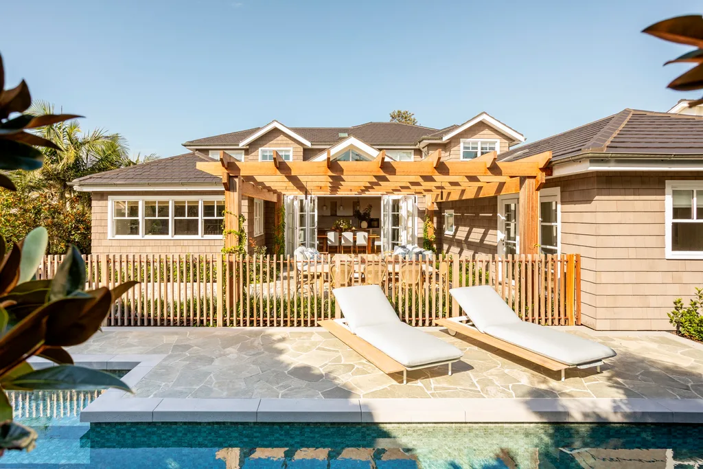 Poolside view of a house with a pergola, patio furniture, and two lounge chairs under a clear blue sky.