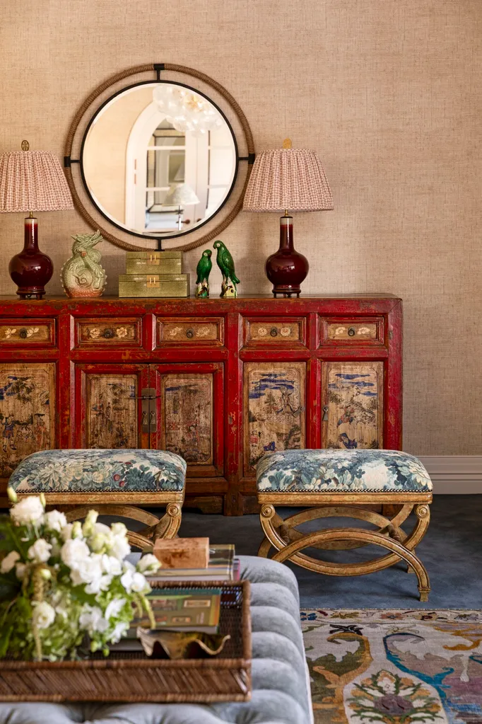 Stylish room with red antique Chinese cabinet, round mirror on the wall above. Two ottomans, decorated with floral upholstery are in front. Two table lamps with pleated lampshades in red and white pattern are positioned on top of the antique cabinet, beside two green bird figurines and other chinoiserie decorative objects.