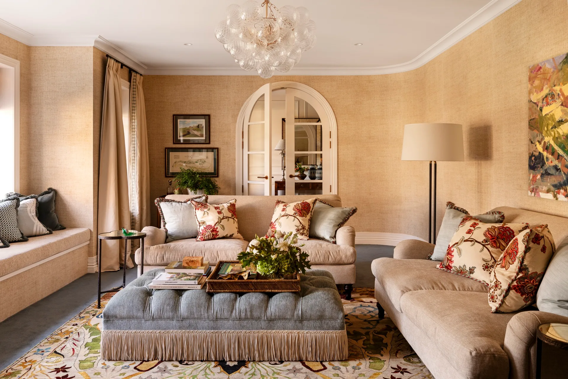Elegant formal sitting room in a Cape Cod style home, with beige sofas, floral pillows, a blue ottoman, chandelier, and arched doors.
