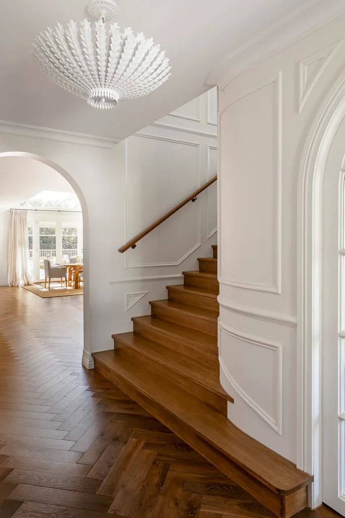 Stylish hallway with wooden staircase, white walls with decorative moulding, ornate chandelier, and an archway leading to a light filled dining space.