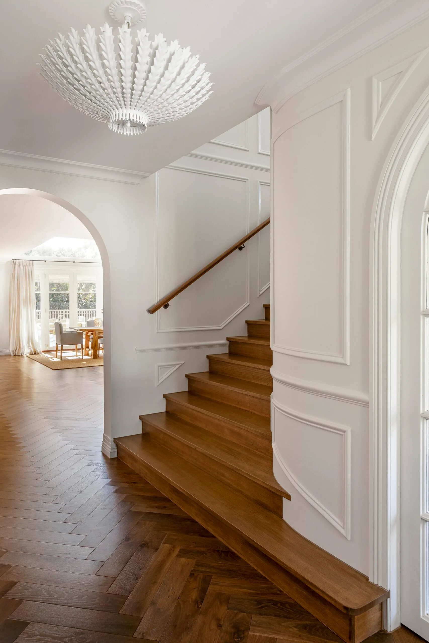 Stylish hallway with wooden staircase, white walls with decorative moulding, ornate chandelier, and an archway leading to a light filled dining space.