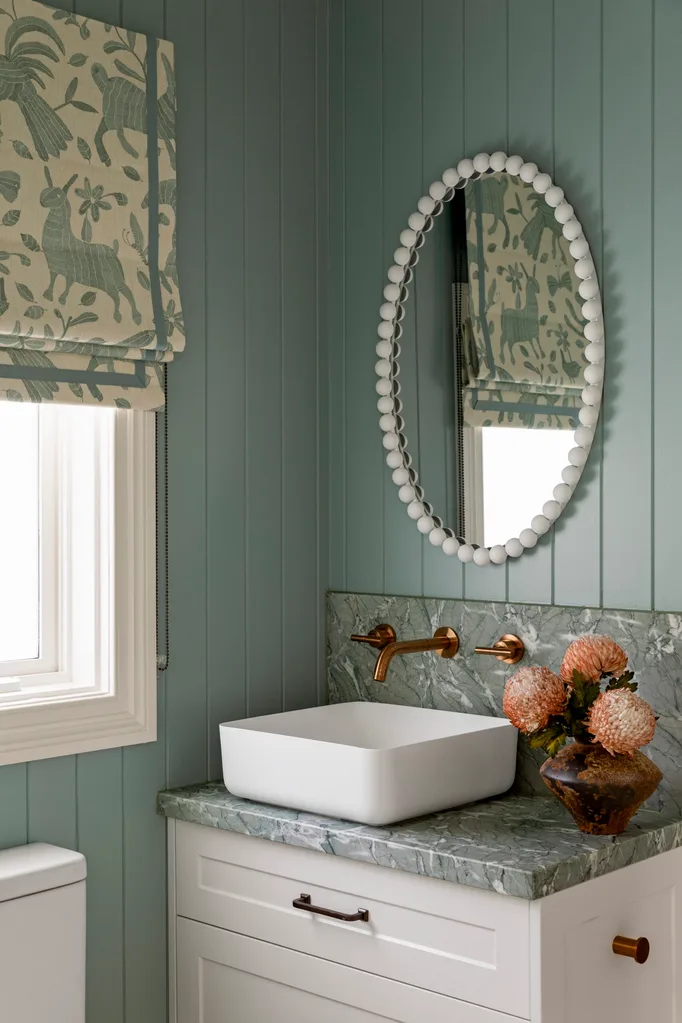 Bathroom with teal panelled walls, teal toned marble splashback and benchtop attached to a white vanity with white sink, all underneath a white framed oval mirror. The hardware is copper and a floral Roman blind, in matching cream and teal, is positioned on a window. This is in a Cape Cod style home.
