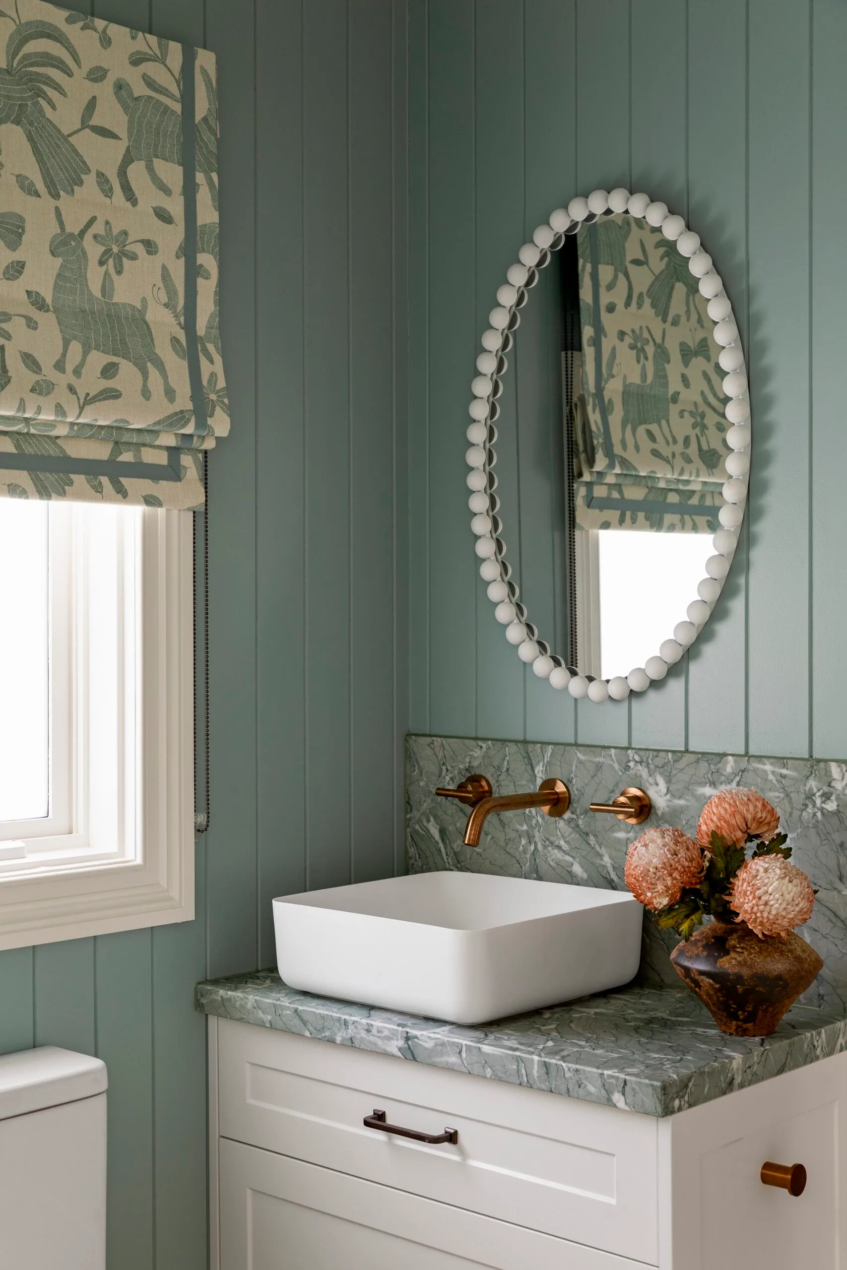 Bathroom with teal panelled walls, teal toned marble splashback and benchtop attached to a white vanity with white sink, all underneath a white framed oval mirror. The hardware is copper and a floral Roman blind, in matching cream and teal, is positioned on a window. This is in a Cape Cod style home.