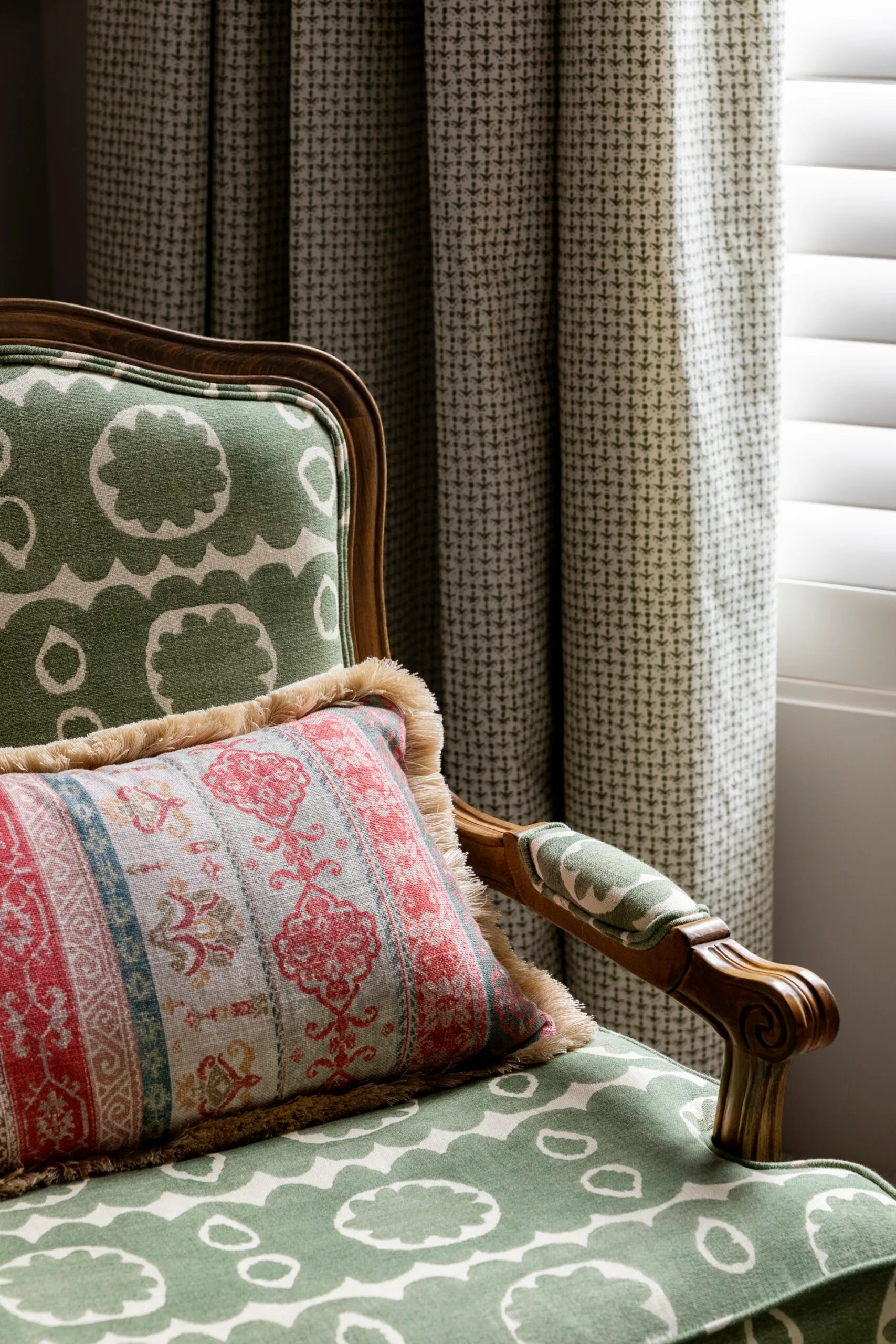 Armchair with green patterned upholstery and a colorful fringed cushion beside patterned curtains.