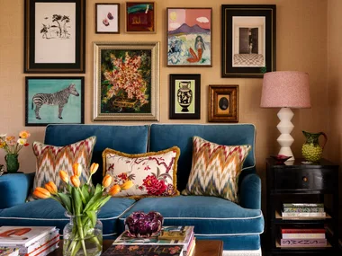 Living room with a blue sofa, colorful cushions, a gallery wall of art, side table with lamp, and tulip vase.