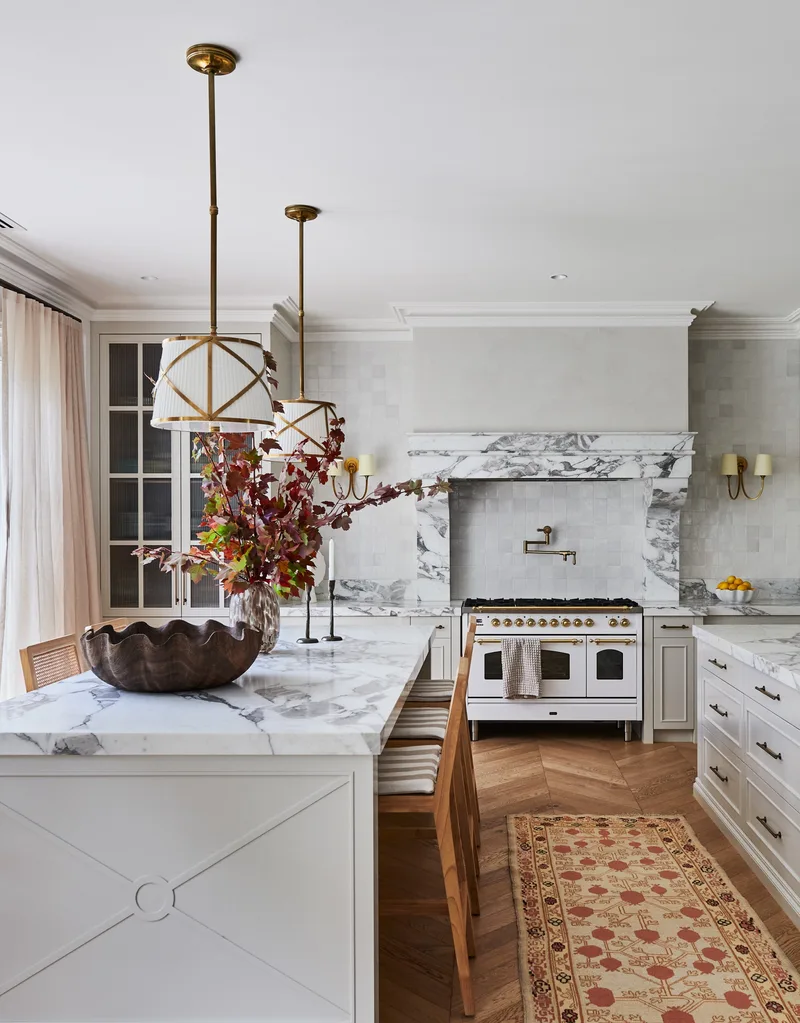 Bright kitchen with marble countertops, wooden stools, a decorative rug, and pendant lights above the island.