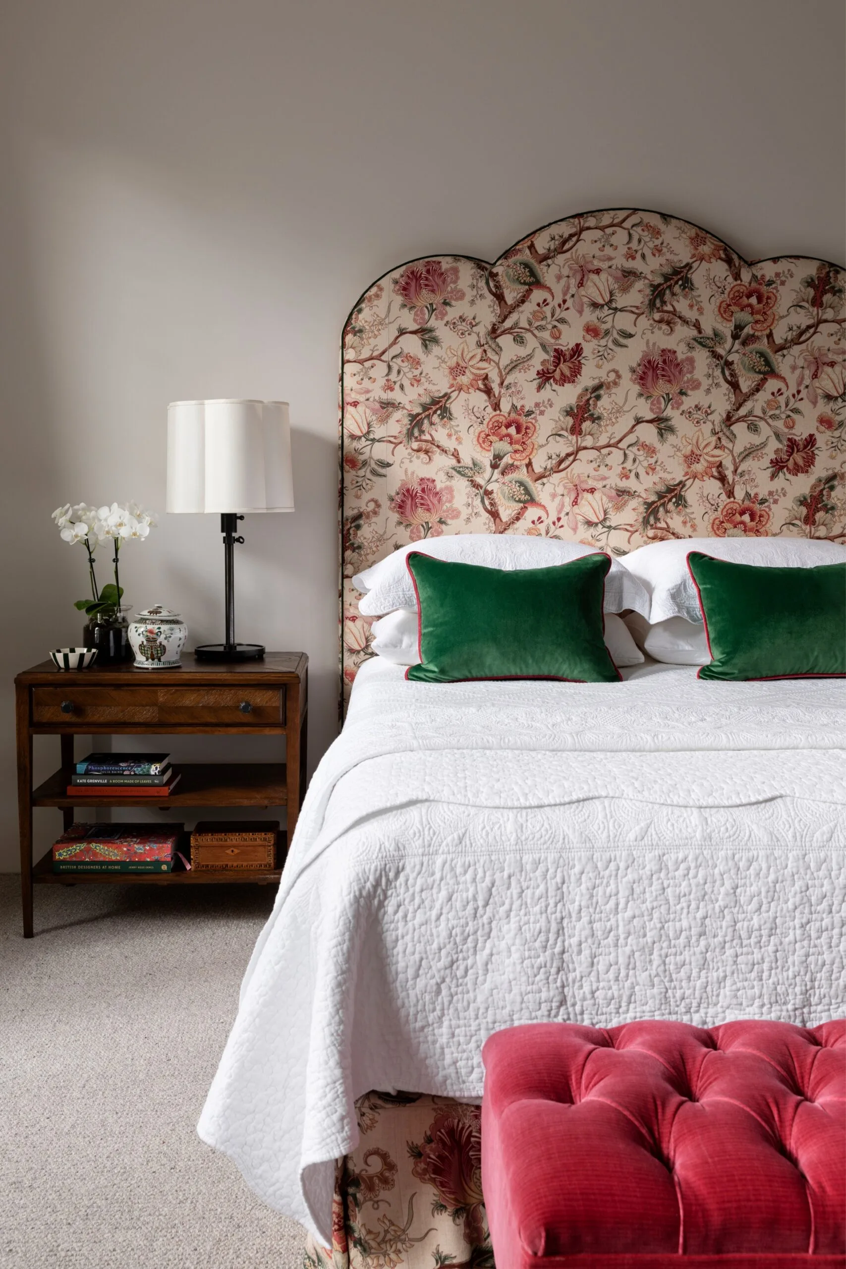 Floral headboard and green pillows on bed with white quilt; wooden nightstand with lamp and books; red ottoman in front.