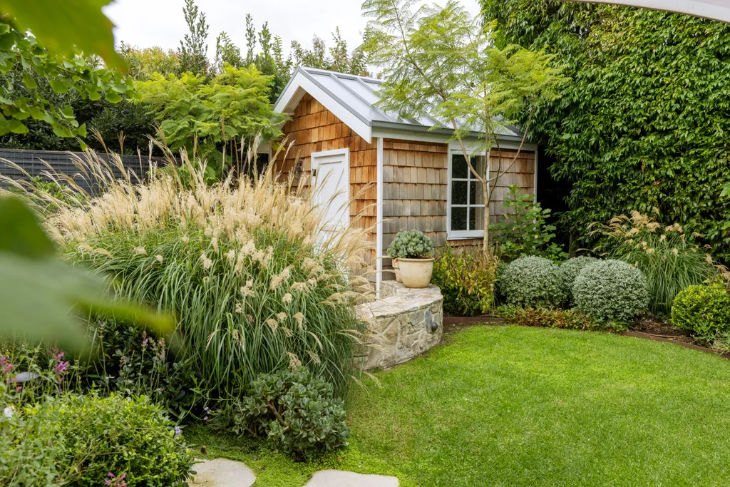 Small rustic garden shed with a shingled roof, surrounded by lush greenery and ornamental grasses, including evergreen miscanthus.