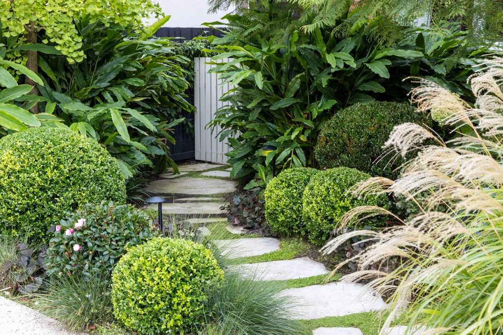 Lush garden path lined with dense green shrubs and ornamental grasses leading to a wooden gate.