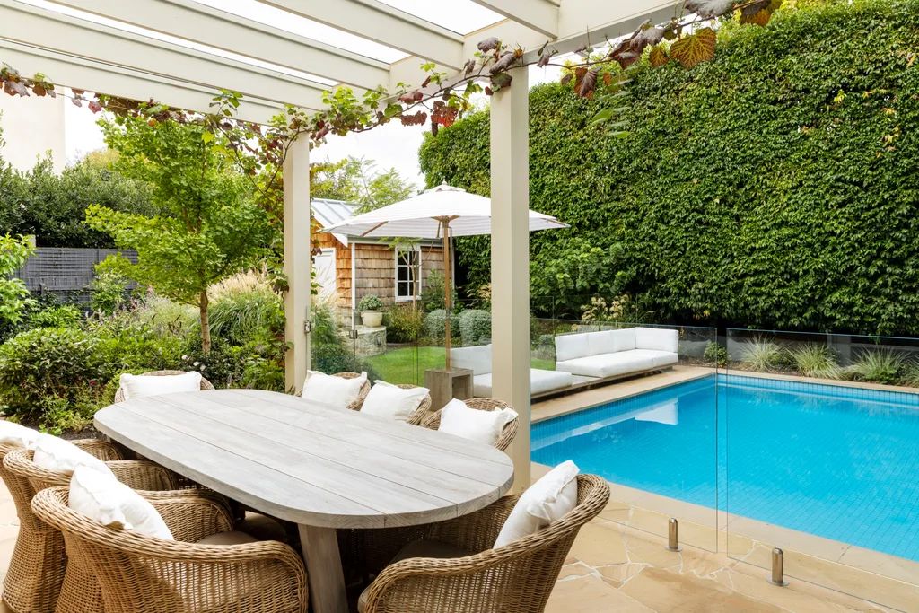 Alfresco dining area with a wicker dining set under a pergola, which has crimson glory vine growing on it. This is next to a swimming pool, surrounded by lush greenery.