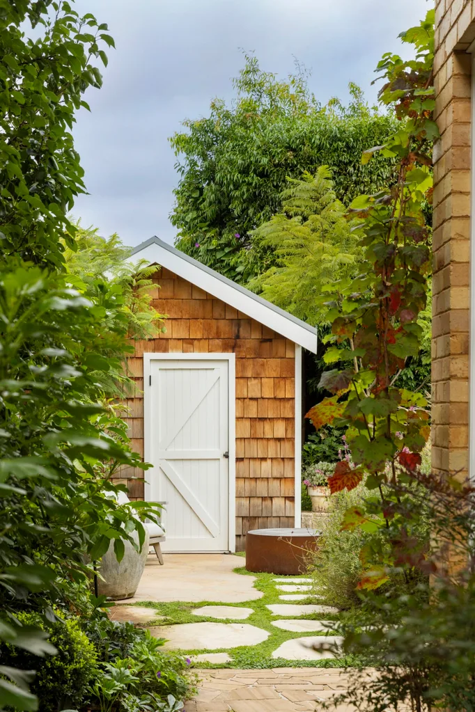A small shingled garden shed with a white door surrounded by lush greenery and stepping stones. Crimson glory vine grows to the right of the image, near a brick wall.