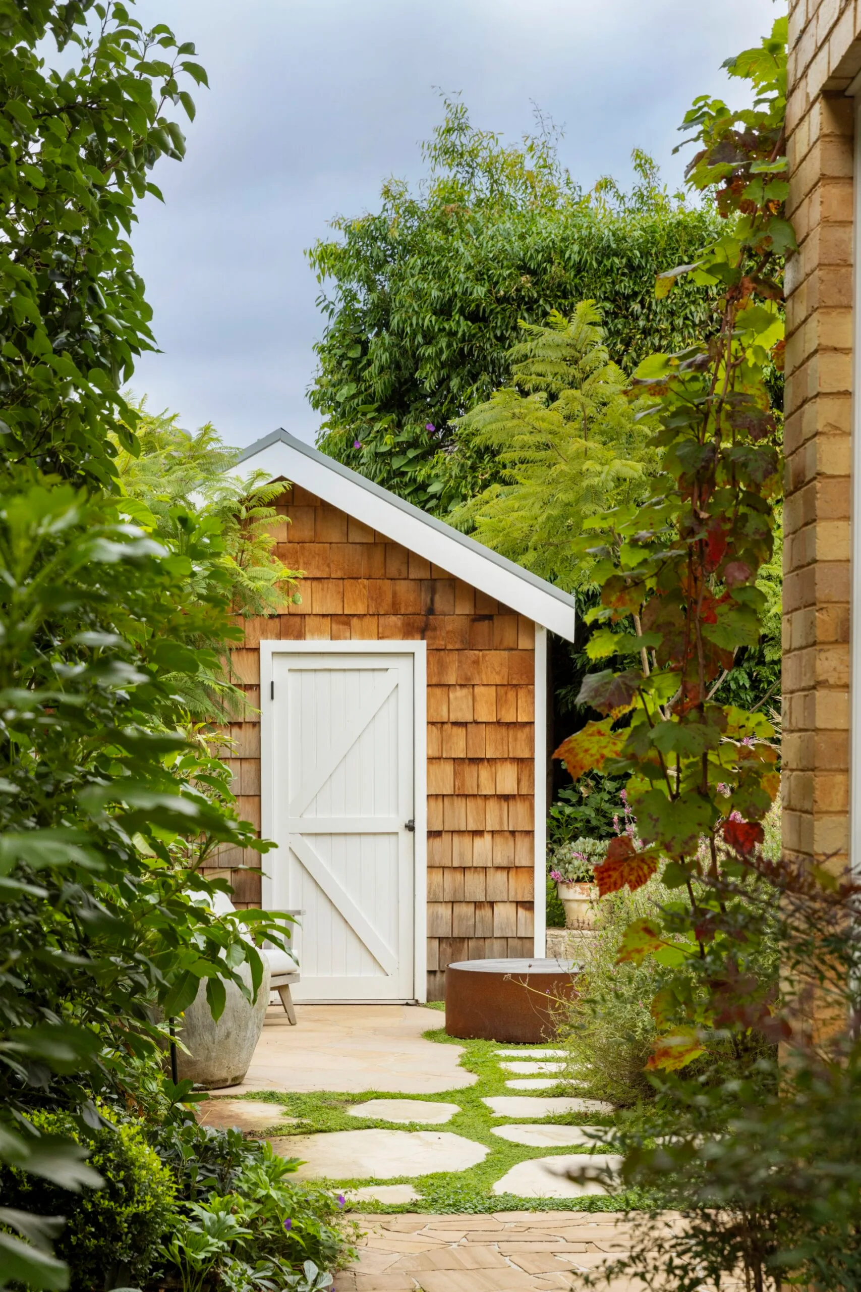 A small shingled garden shed with a white door surrounded by lush greenery and stepping stones. Crimson glory vine grows to the right of the image, near a brick wall.