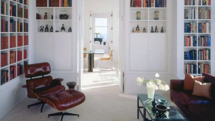 An eames chair sits to one side of a library with white walls and inbuilt bookshelves lined with books