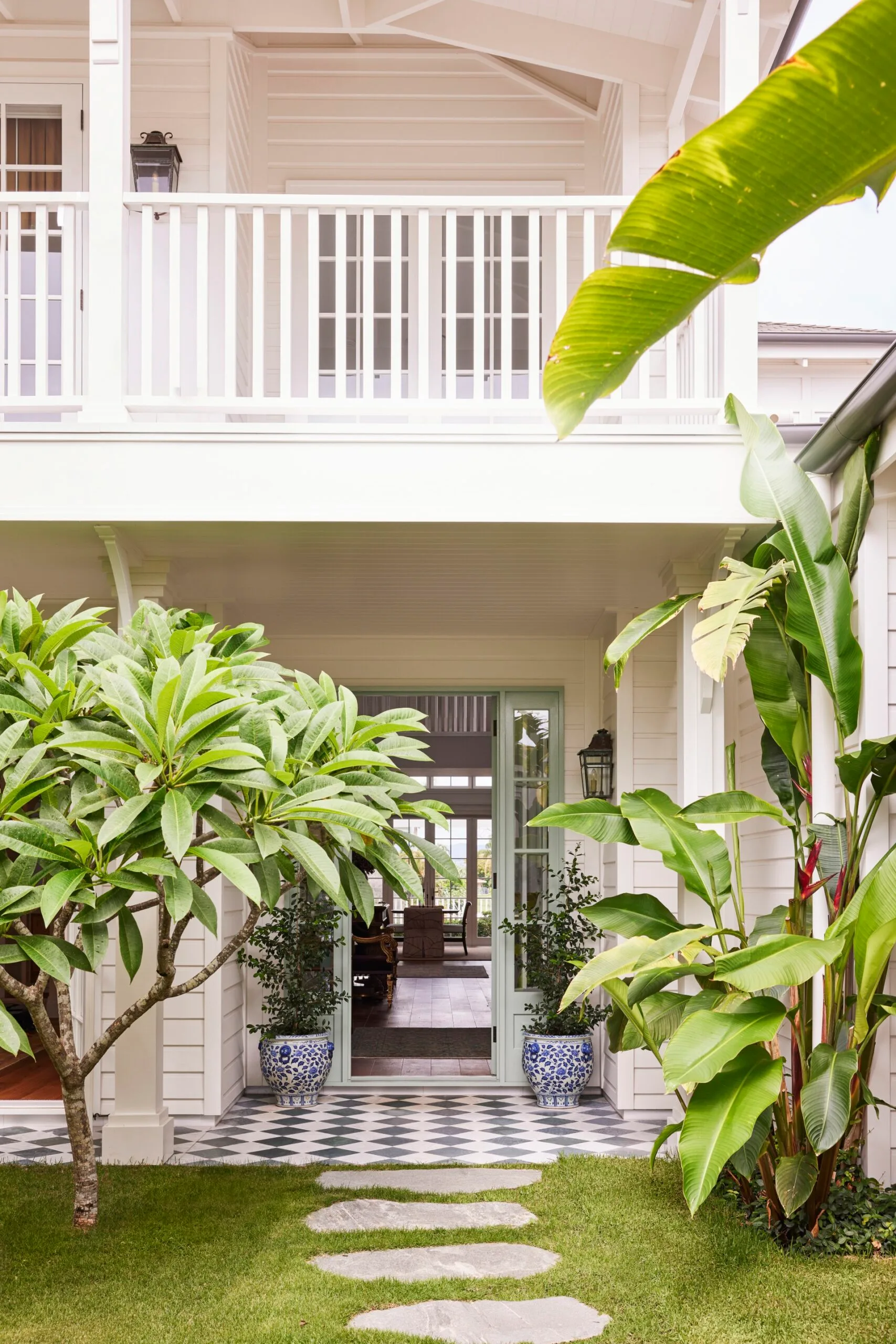 White house with checkered tile entrance, green plants, and balcony above. Pathway of stone slabs leads to door.