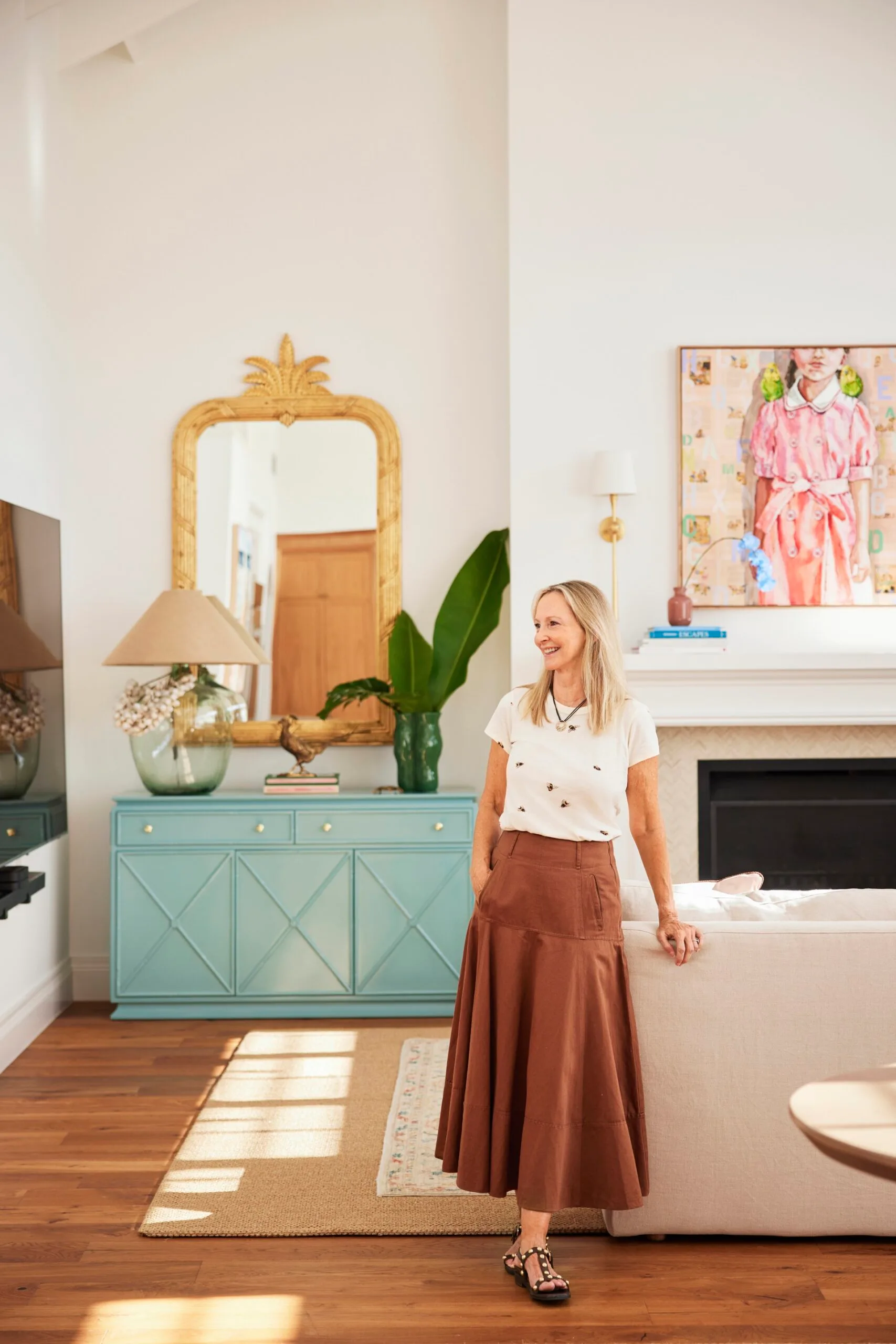 A woman in a white shirt and brown skirt smiles, standing in a stylish, well-decorated living room.