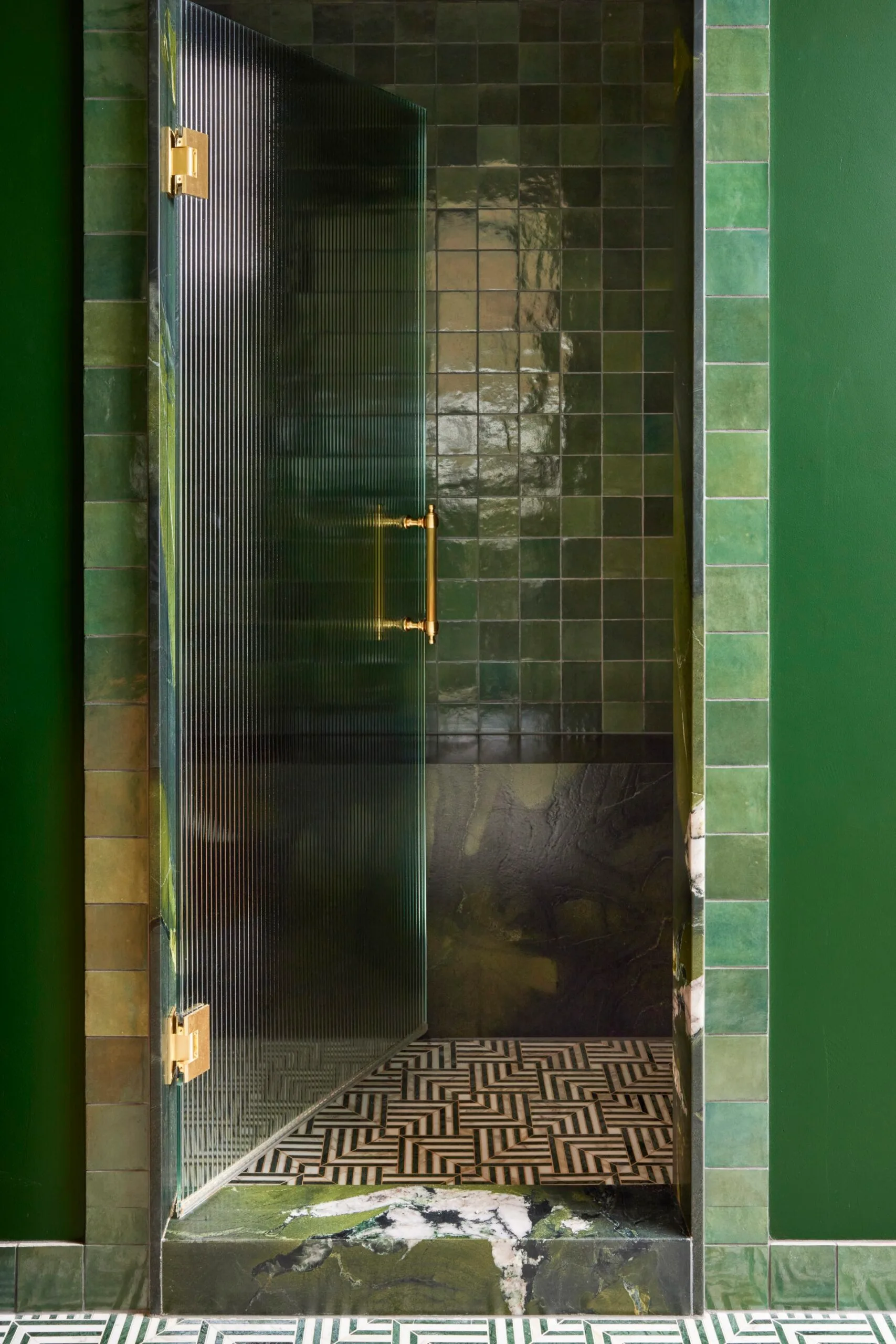 Green tiled shower with a glass door and geometric patterned floor in a bathroom.