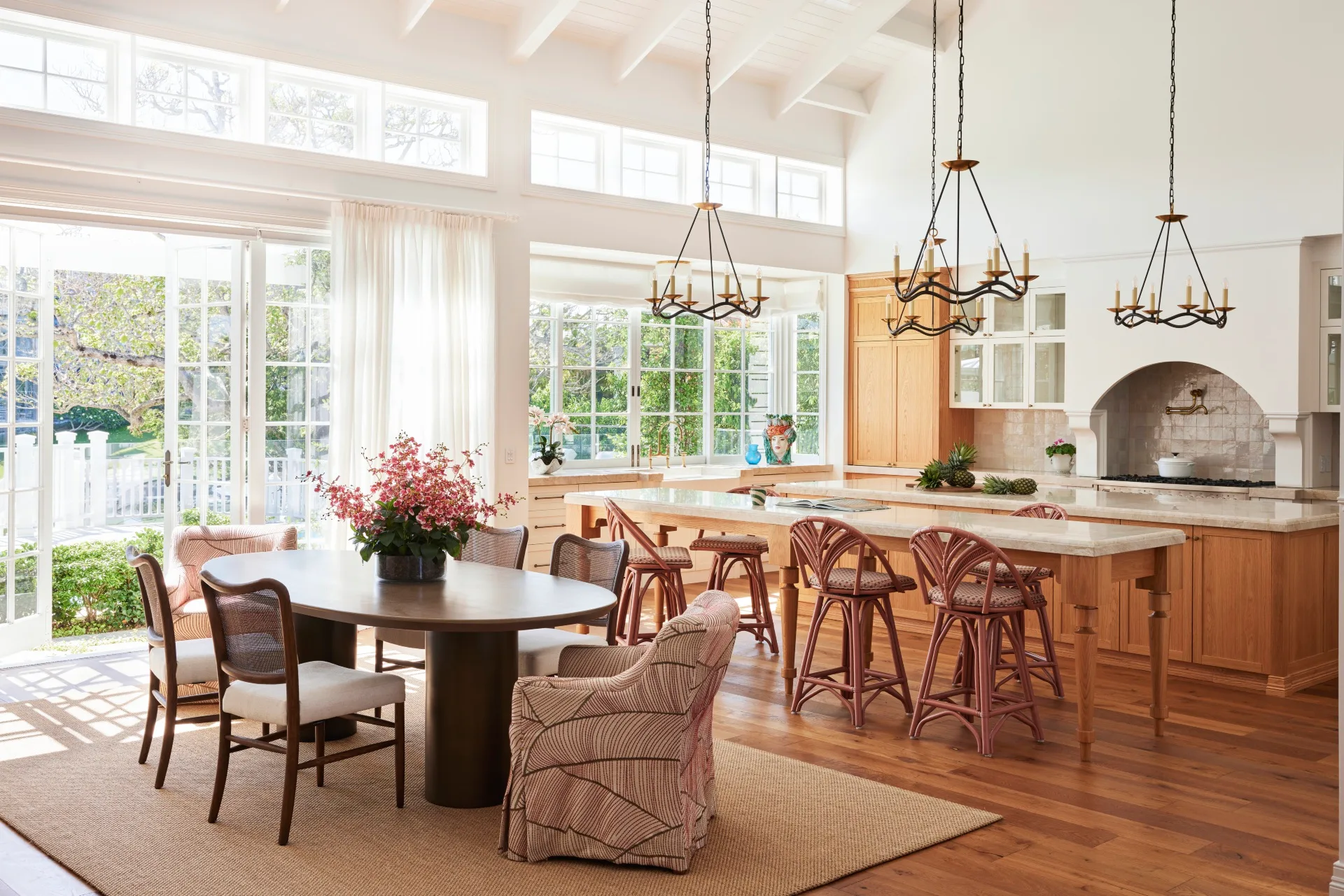 Bright, spacious kitchen with wooden cabinets, large windows, chandeliers, and a round dining table with flowers.