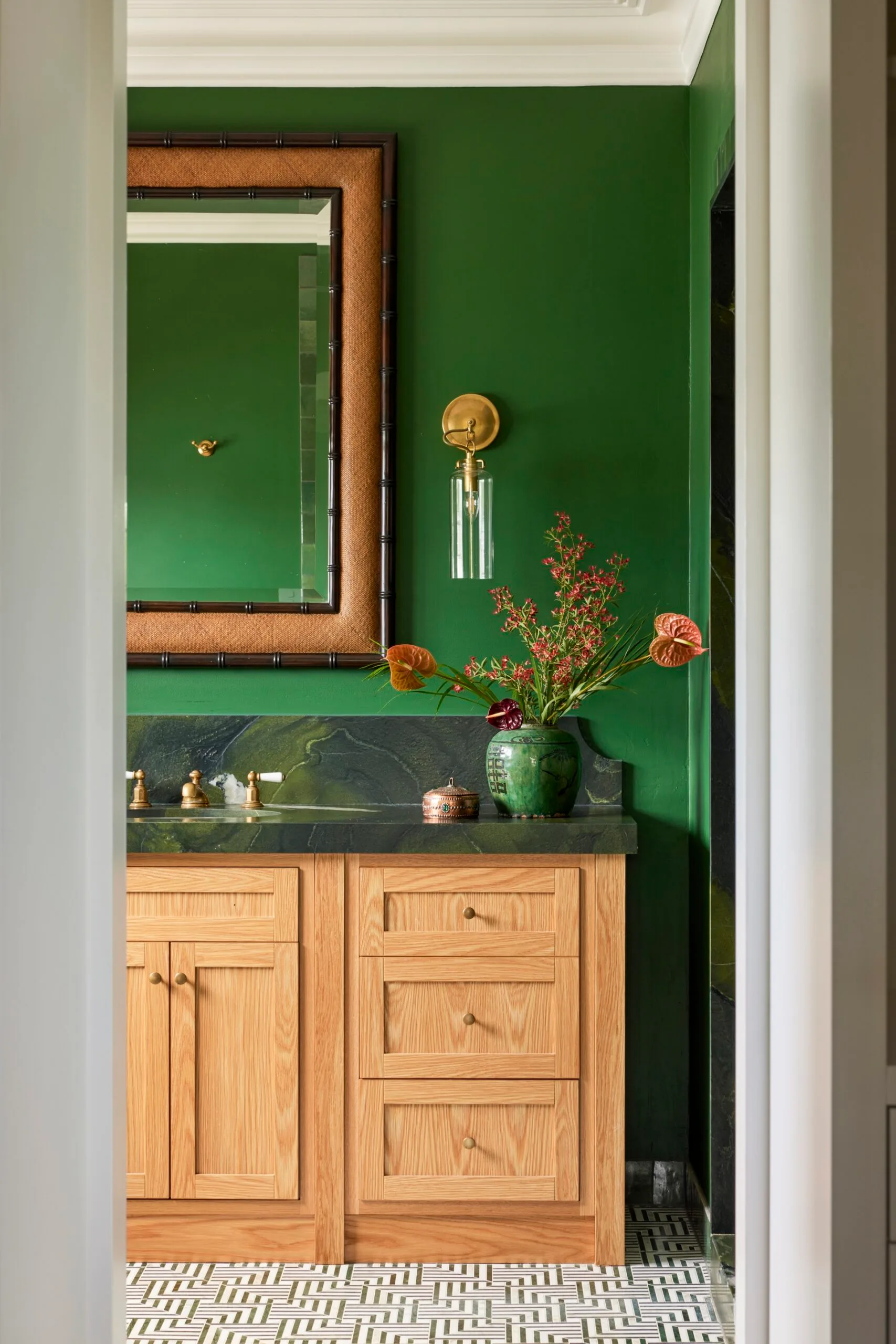 Green bathroom with wooden vanity, gold fixtures, large mirror, and floral vase on marble counter.