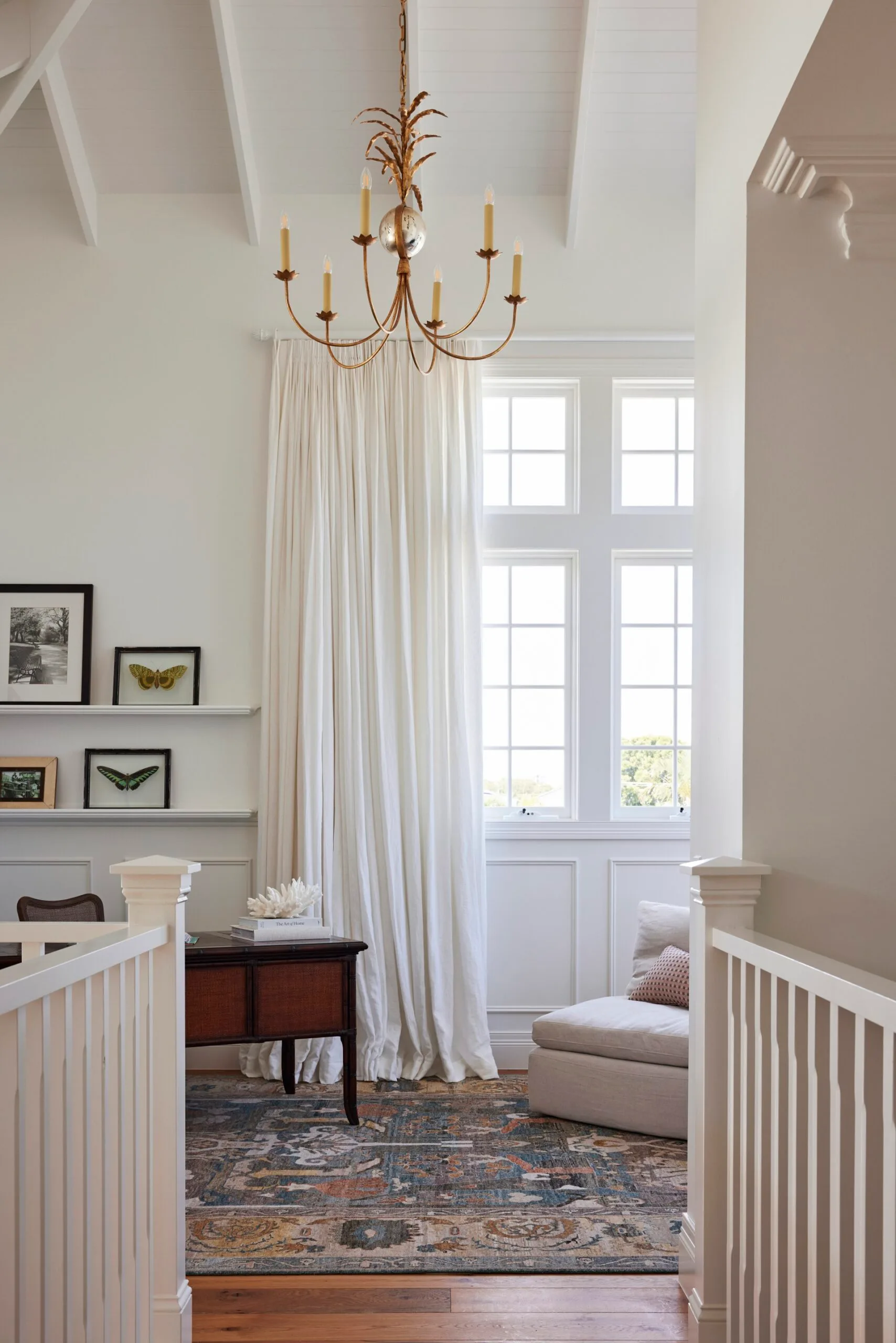 Bright room with high windows, white curtains, a chandelier, framed butterfly art, a wooden table, and a patterned rug.