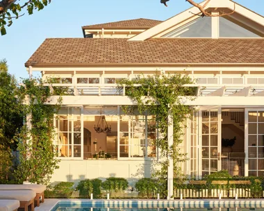 White house with large windows, greenery, and a pool reflecting the building against a clear blue sky.