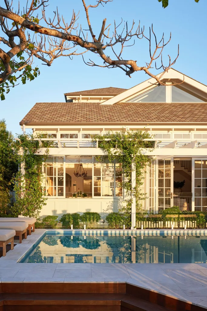 White house with large windows, greenery, and a pool reflecting the building against a clear blue sky.