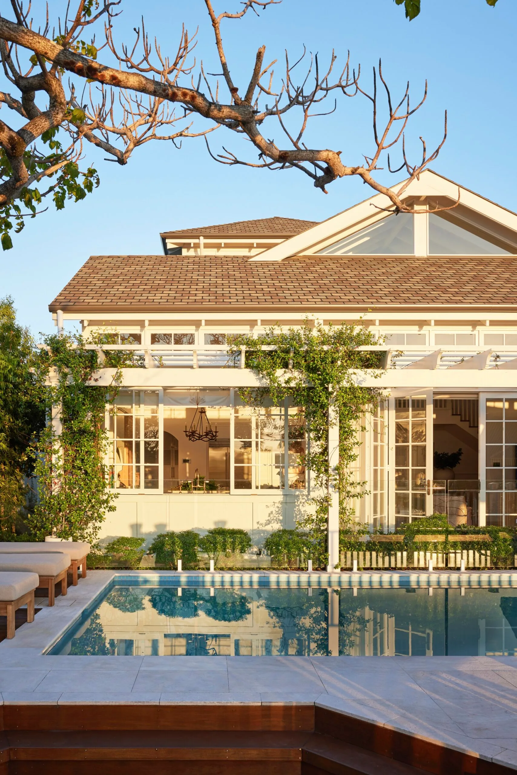 White house with large windows, greenery, and a pool reflecting the building against a clear blue sky.