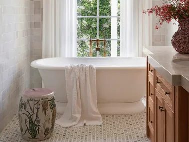 Elegant bathroom with a freestanding tub, white curtains, floral arrangement, tiled floor, and natural light from a window.