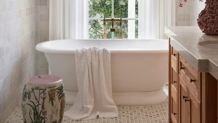 Elegant bathroom with a freestanding tub, white curtains, floral arrangement, tiled floor, and natural light from a window.