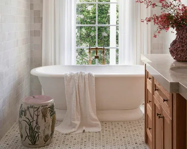 Elegant bathroom with a freestanding tub, white curtains, floral arrangement, tiled floor, and natural light from a window.