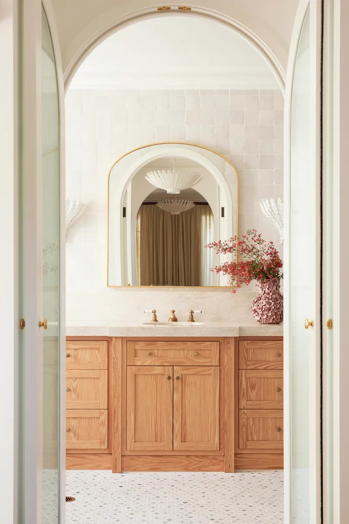 Elegant bathroom with wooden cabinets, arched mirror, gold faucet, chandelier, and pink flowers in a textured vase.