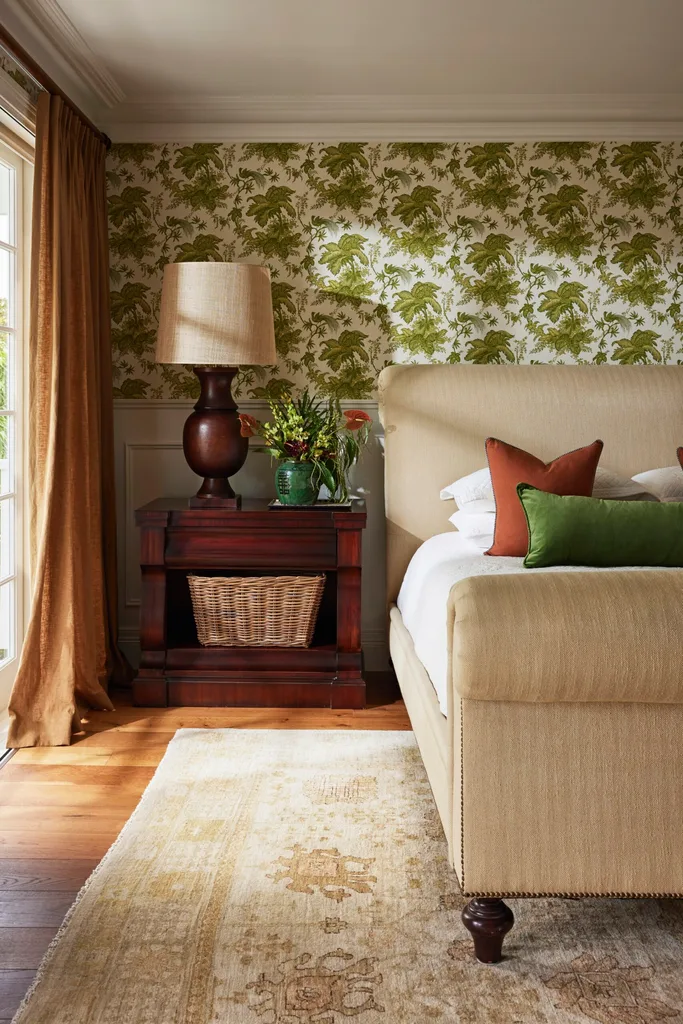 Bedroom with a beige bed, floral wallpaper, wooden side table, wicker basket, lamp, and floral arrangement.